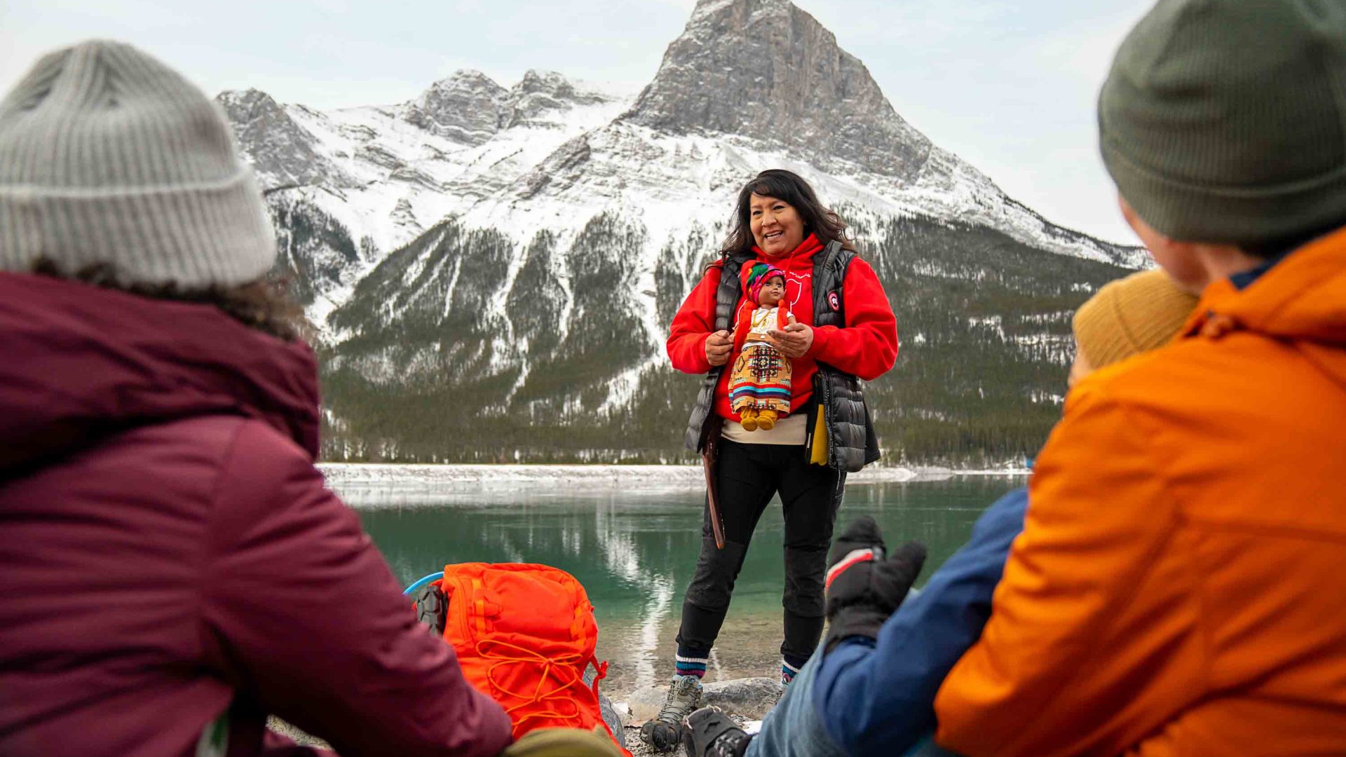 A woman stands in front of mountains and a lake and talks to some travellers who are seated.