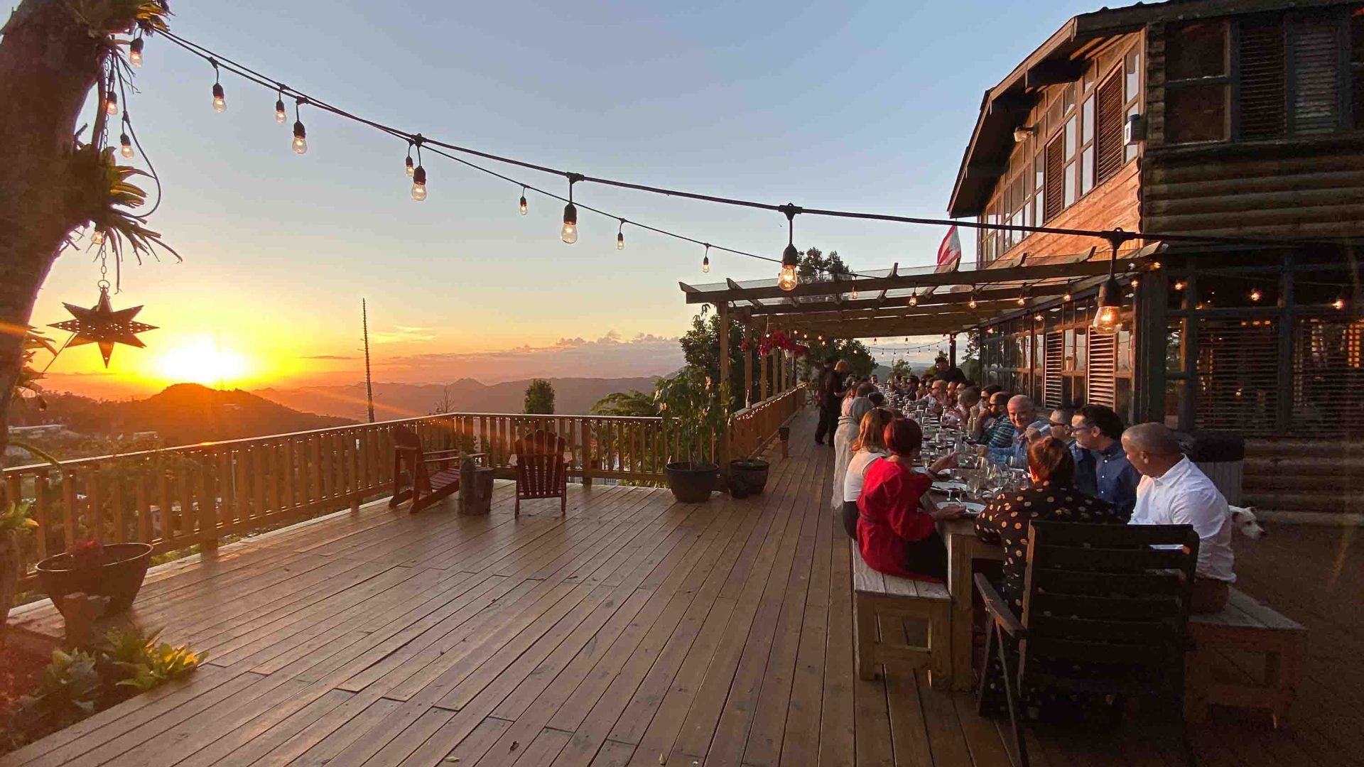 Tourists sit at a long dinner table on a deck overlooking hills and the setting sun.