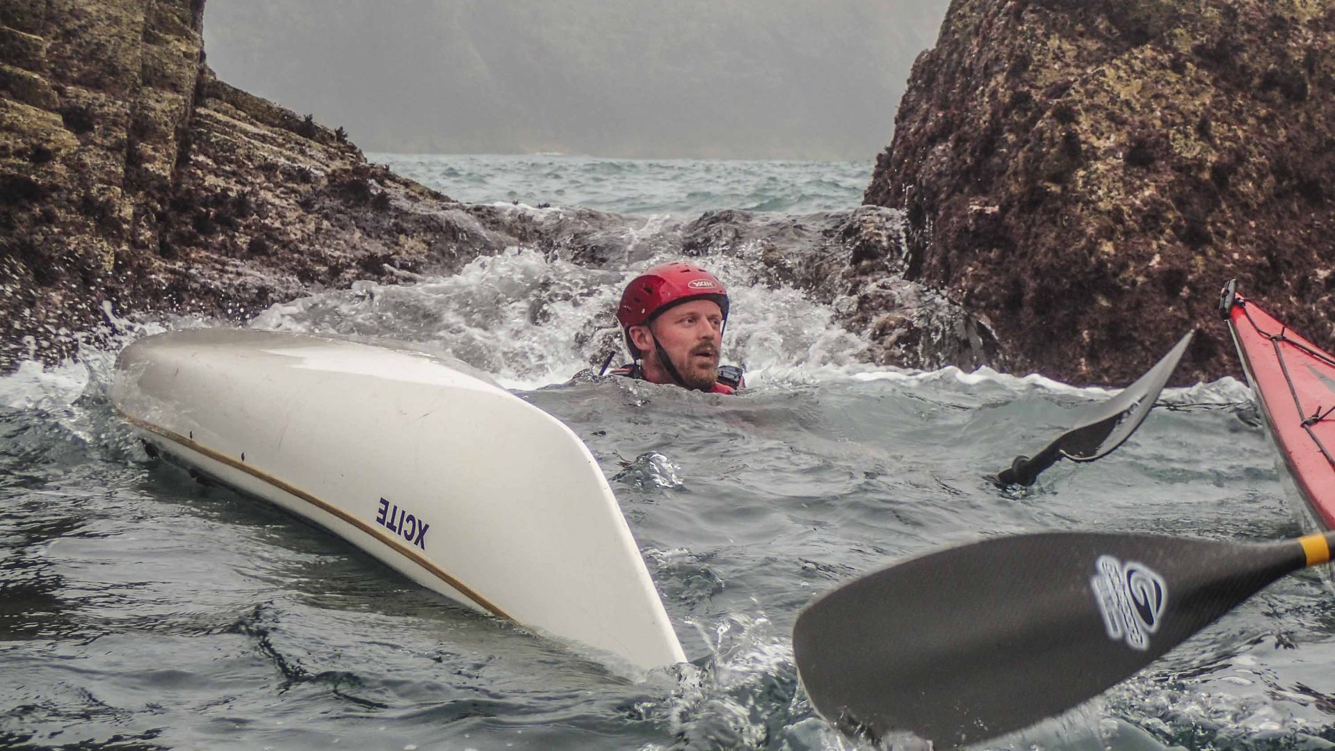 A man in a helmut in the white wash of the water. His kayak is alongside him.