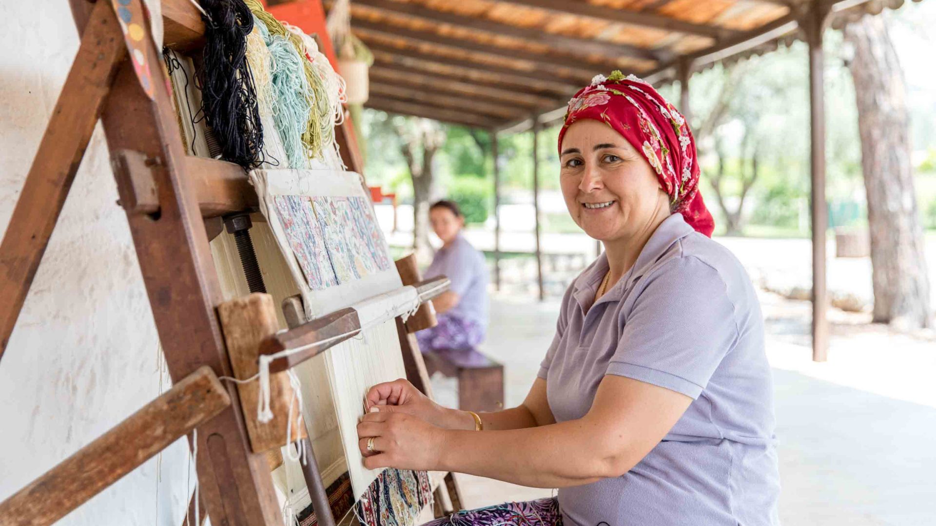 A woman in a red head scarf weaves while seated.